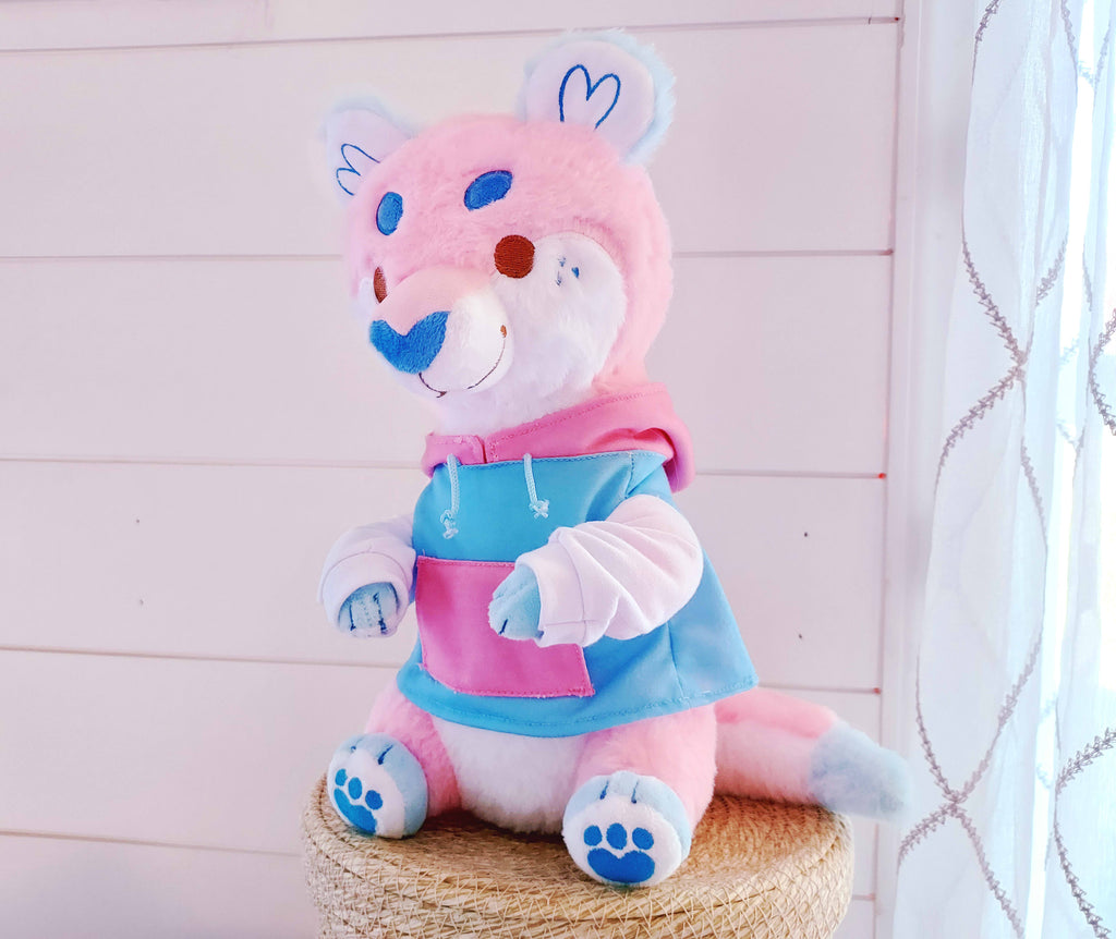 Plush toy of Cake Pop the otter who is pink, white, and blue and wearing a hoodie 