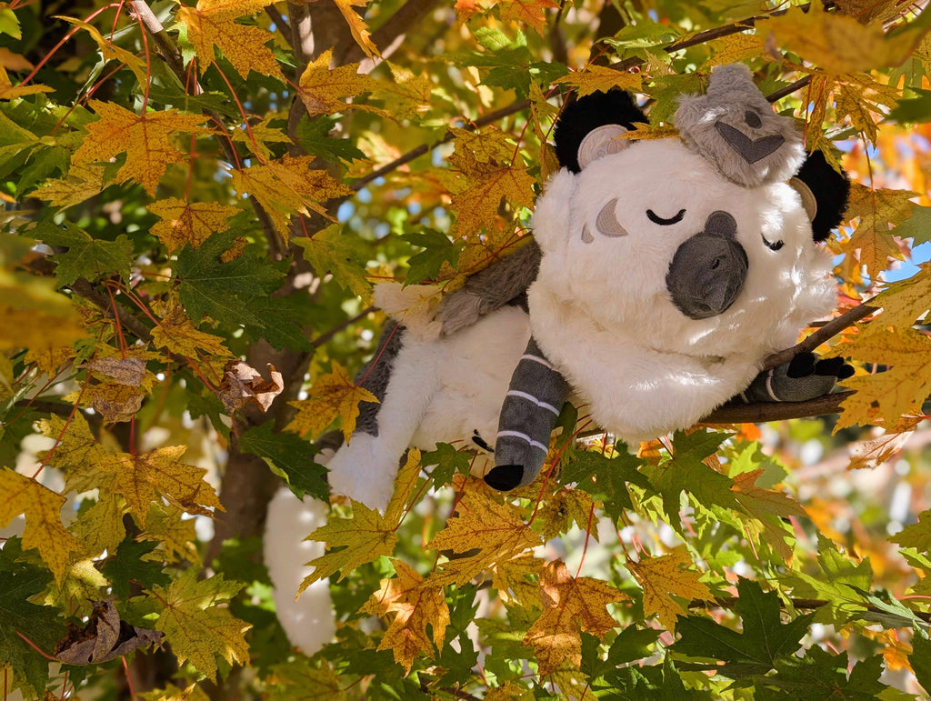 Weighted plush toy of Sesame the grey and white gryphon with his eyes closed, lying in a tree with yellow leaves
