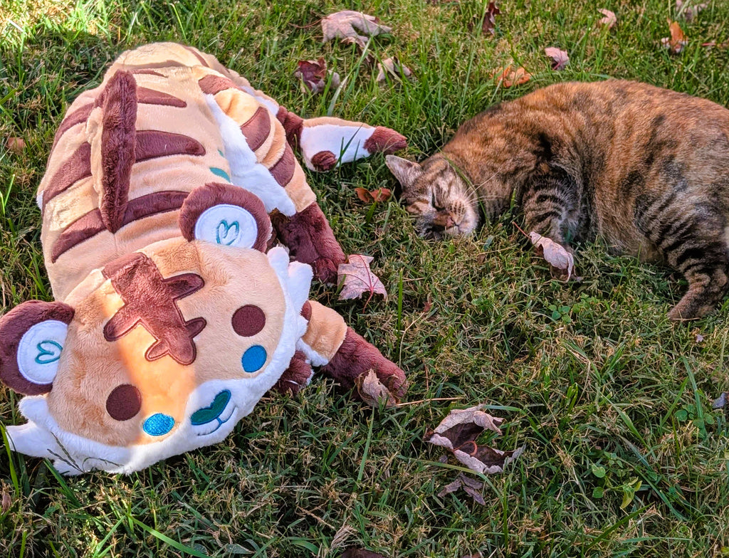 Plush toy of Cornflakes the Tiger and Shark hybrid lying in the grass next to a cat