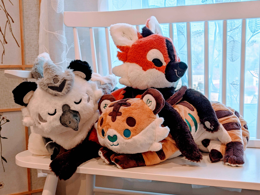 Plush toys of Paprika the maned wolf, Cornflakes the tiger and shark hybrid, and Sesame the grey and white gryphon, lying in a pile on a bench