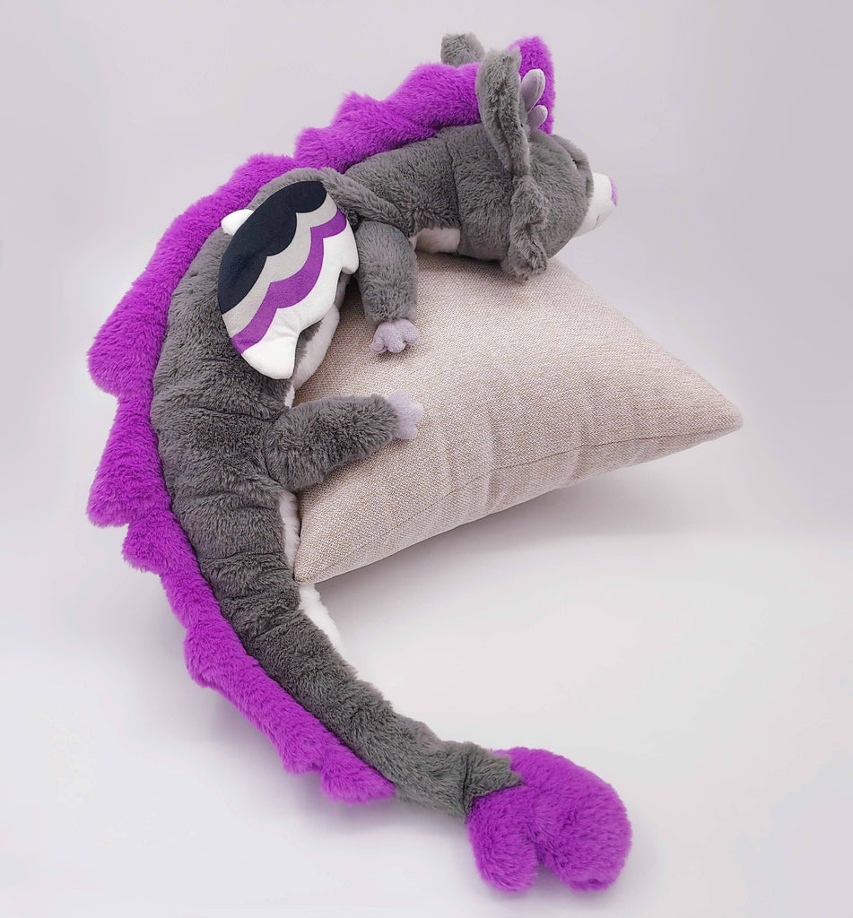 Back view of a large plush of Marshmallow the purple, black, white, and grey dragon, sleeping on a pillow.