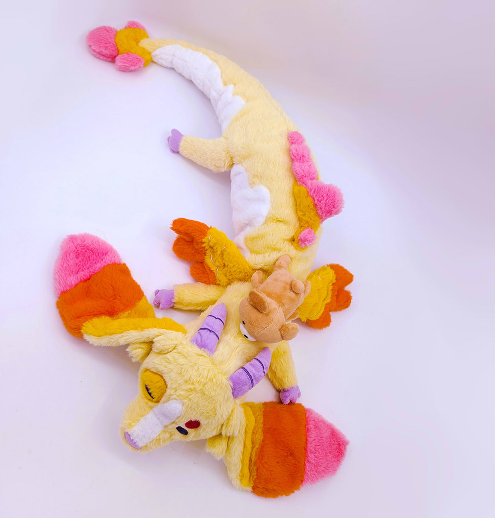 Aeriel view of pillow plush toy of Charmy the dragon who is pink, orange,yellow and has a brown bear toy on his head