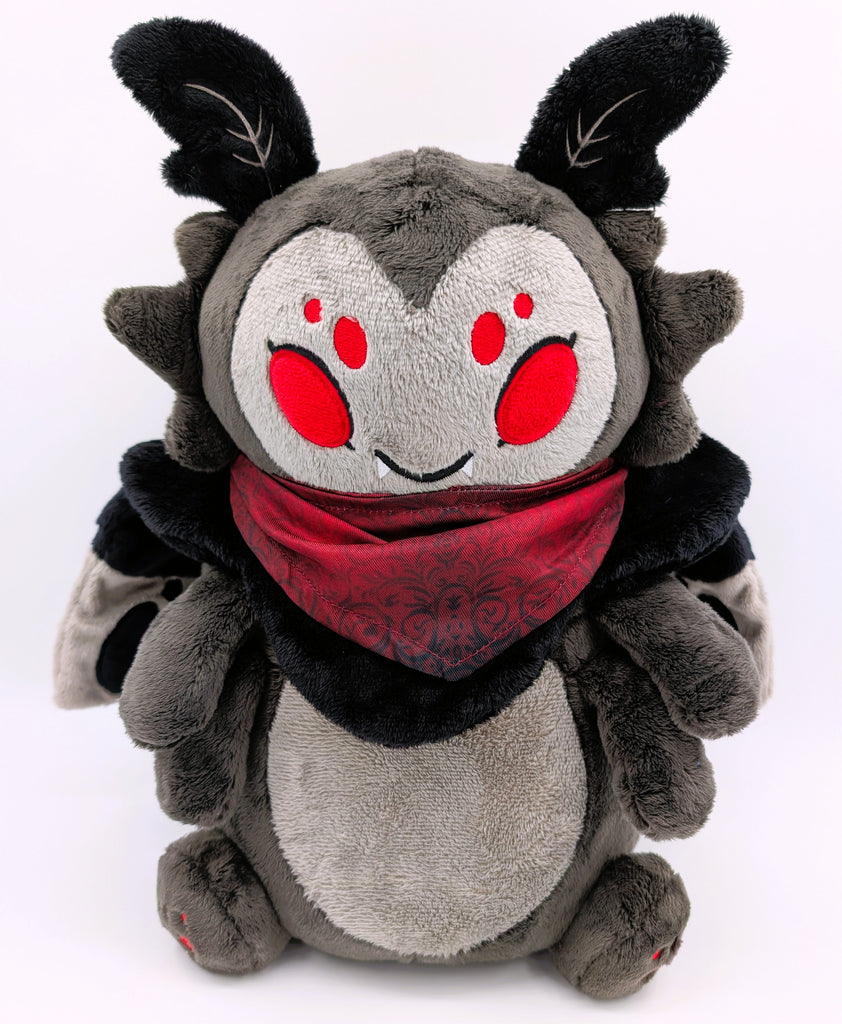 A plush toy of a black moth with red eyes wearing a red bandana. He is smiling and has fangs