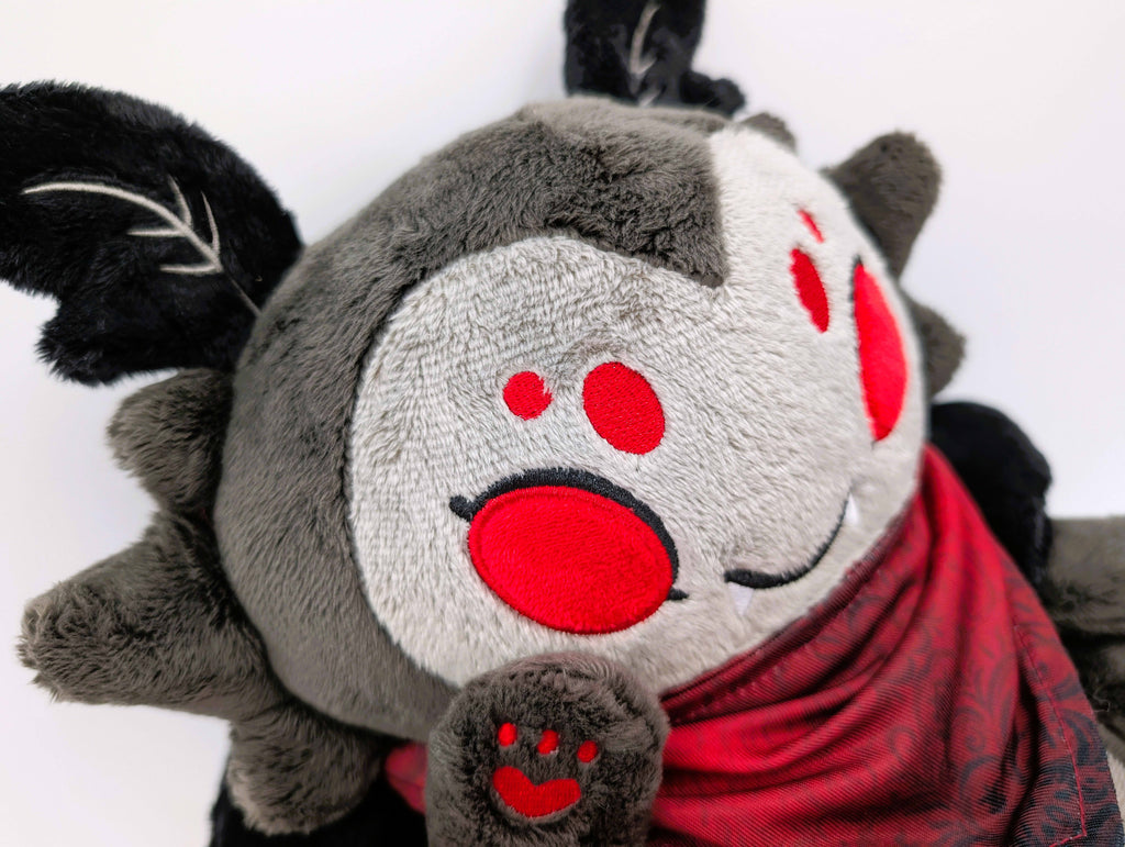 A plush toy of a black moth with red eyes wearing a red bandana. He is smiling and has fangs while waving at the camera