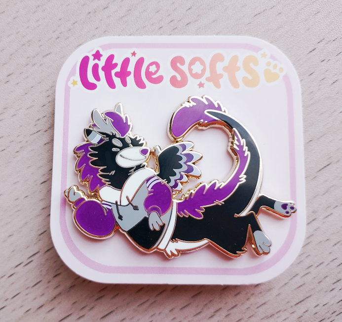 Enamel pin of Marshmallow the dragon. He is black, grey, white, and purple and wearing a hoodie in those same colors.