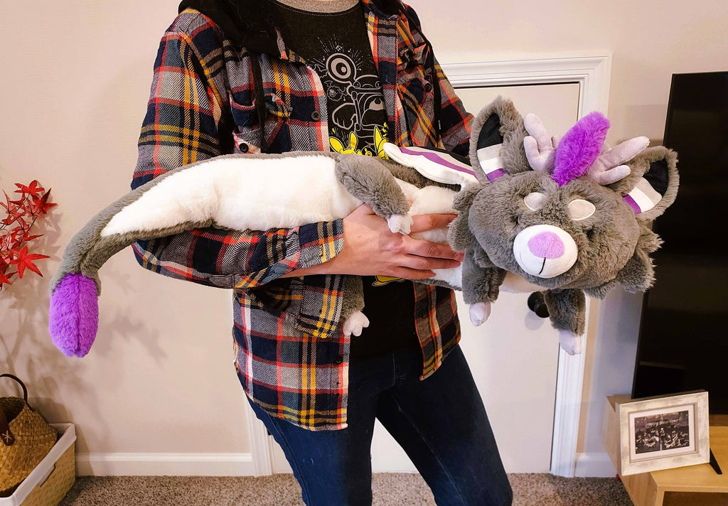 A person holding a large plush of Marshmallow the purple, black, white, and grey dragon. He is asleep.