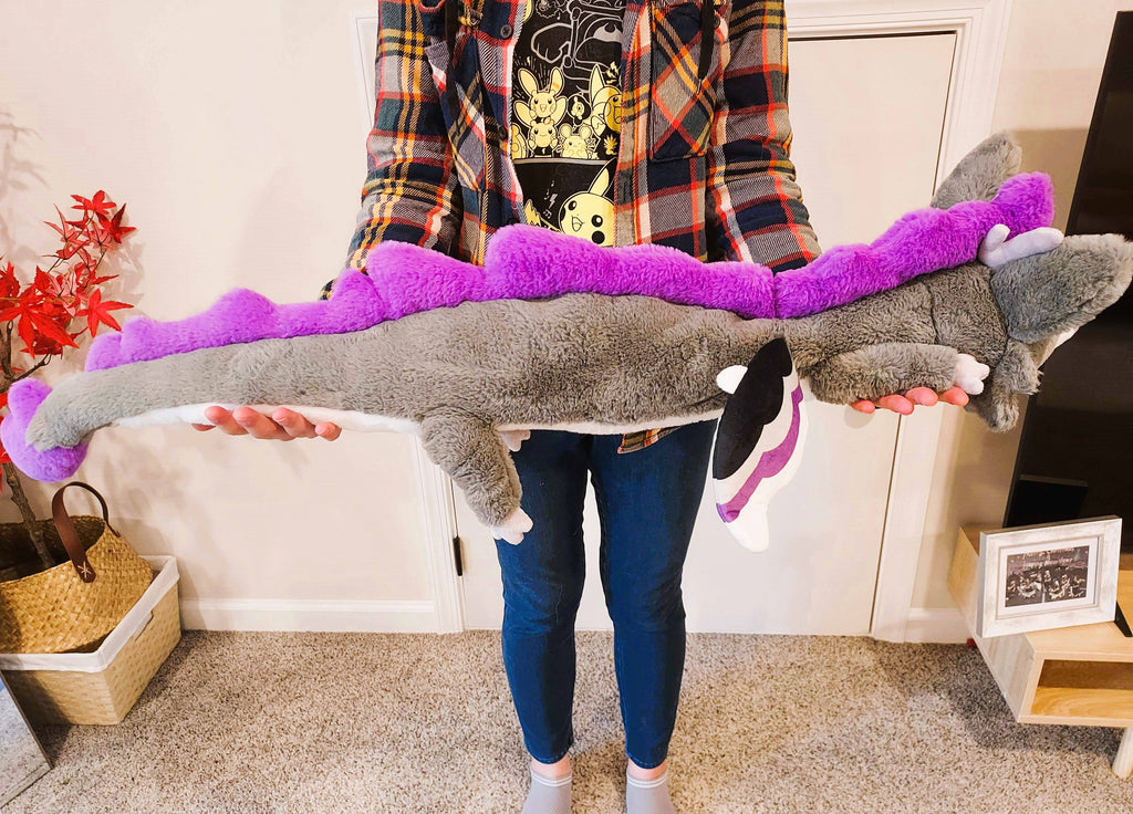 A person holding a large plush of Marshmallow the purple, black, white, and grey dragon. He is asleep.