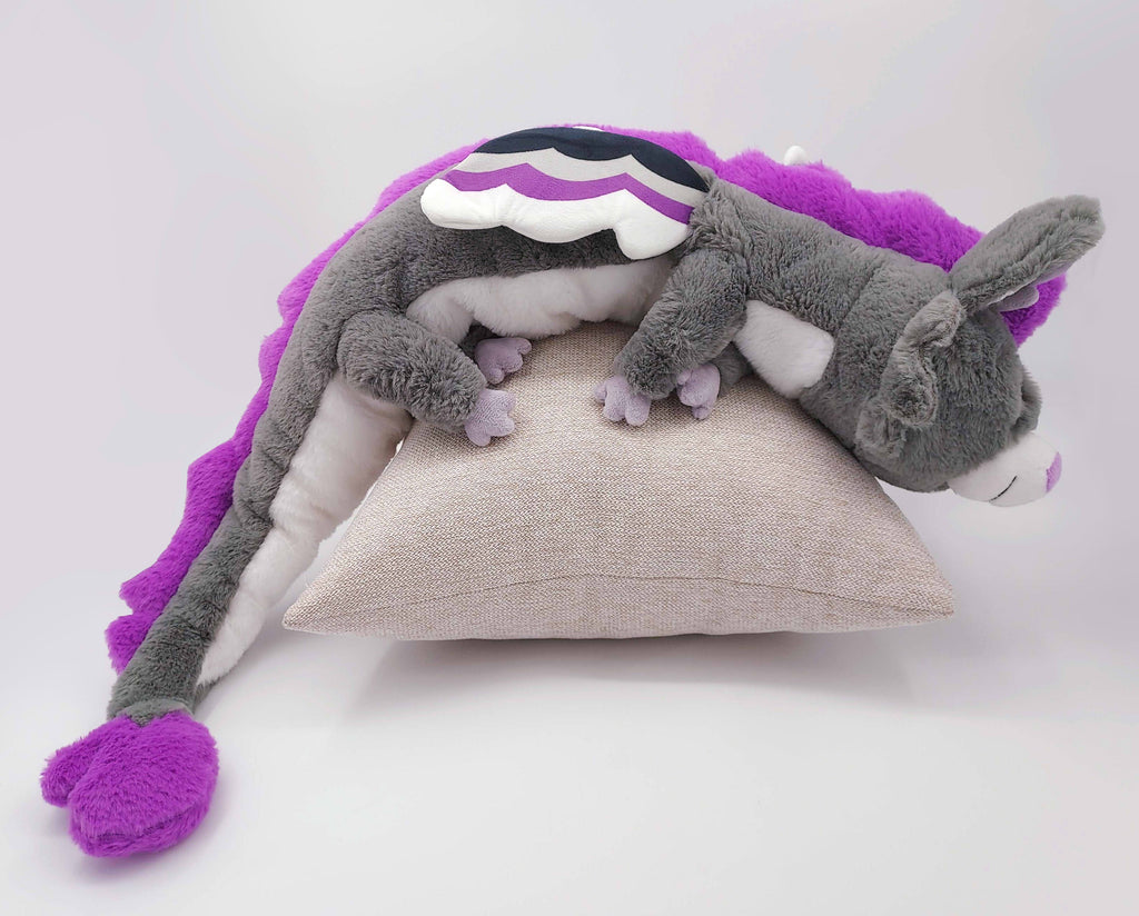 Large plush of Marshmallow the purple, black, white, and grey dragon. He is asleep on a pillow. 