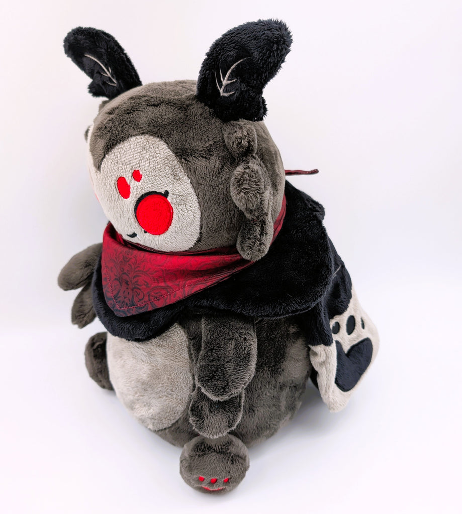 Side view of a plush toy of a black moth with red eyes wearing a red bandana. He is smiling and has fangs