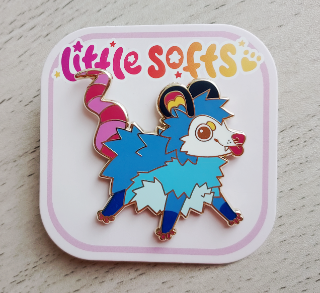 Enamel pin of Pancake the possum. They are blue, pink, and yellow.