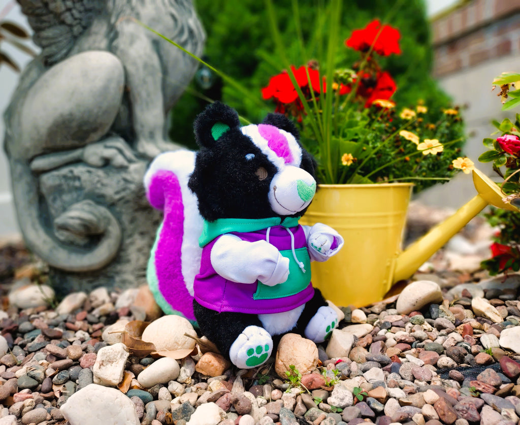 Plush toy of Pepper the green, purple, and white skunk, wearing a matching hoodie sitting near a yellow watering can with red flowers