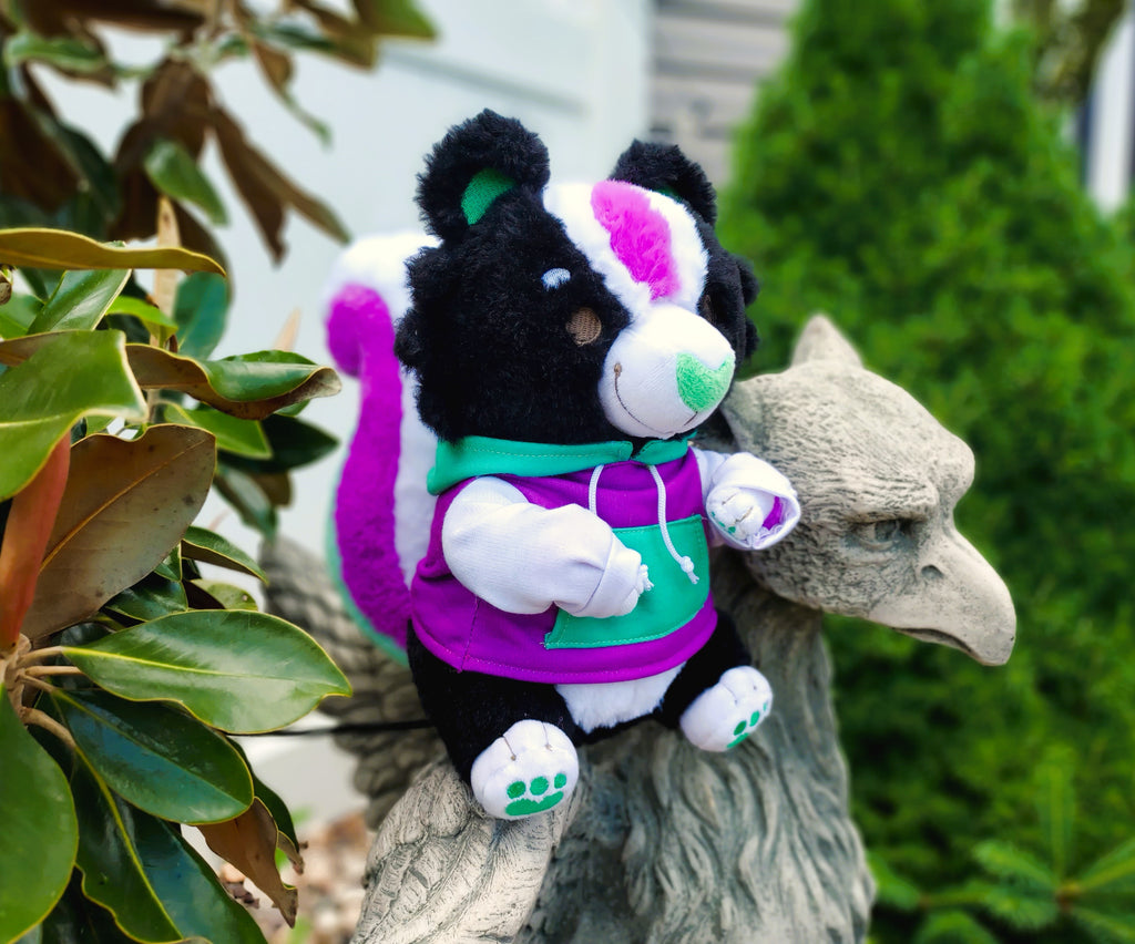 Plush toy of Pepper the green, purple, and white skunk, wearing a matching hoodie and sitting on a gryphon statue