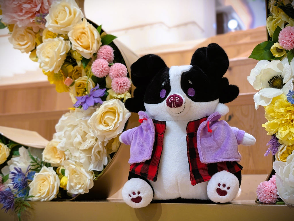 CosyPaws plush of Starry Pup the black and white border collie wearing a purple jacket and sitting next to flowers