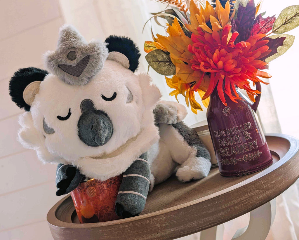 Plush toy of Sesame the grey and white gryphon on a table next to potted flowers