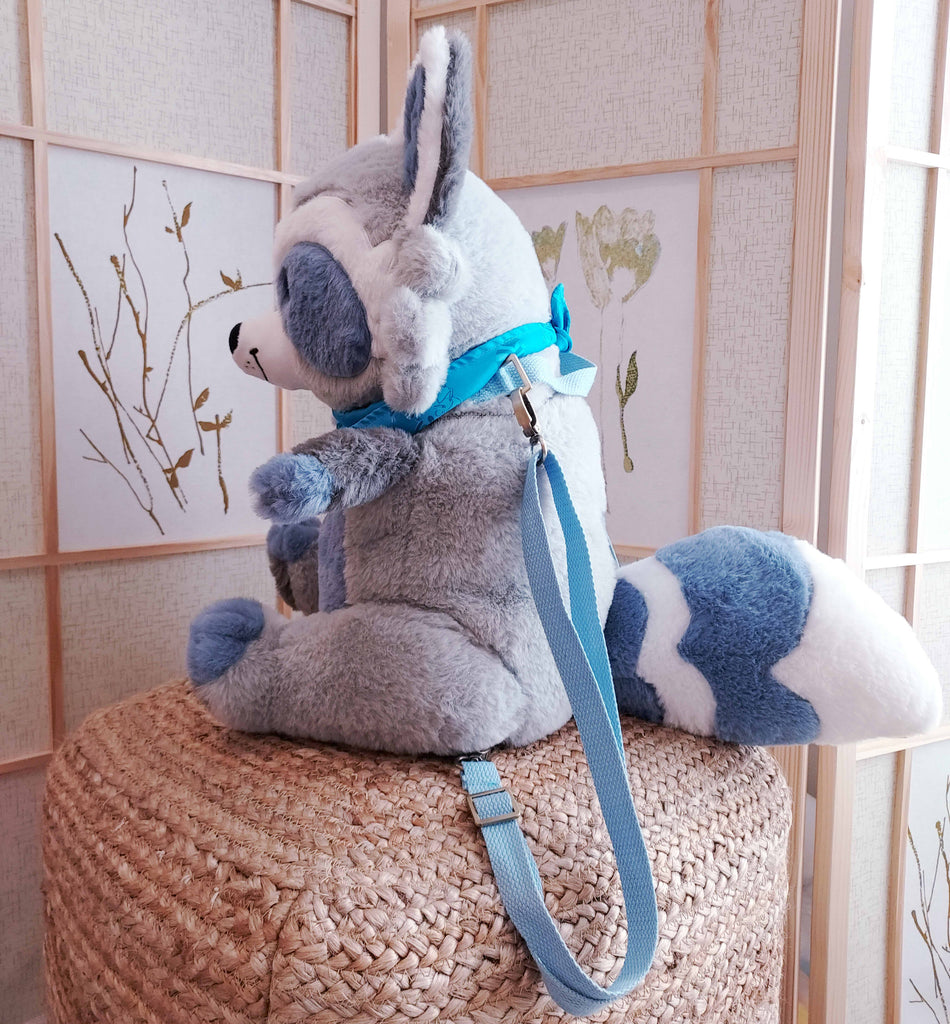 Side view of a plush backpack of Peanut the raccoon wearing a blue bandana, with blue backpack straps