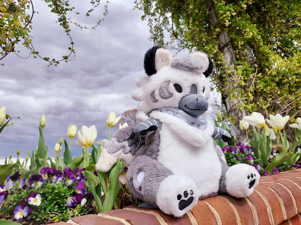 Plush backpack of Sesame the grey and white gryphon sitting on a brick wall by some purple and white flowers