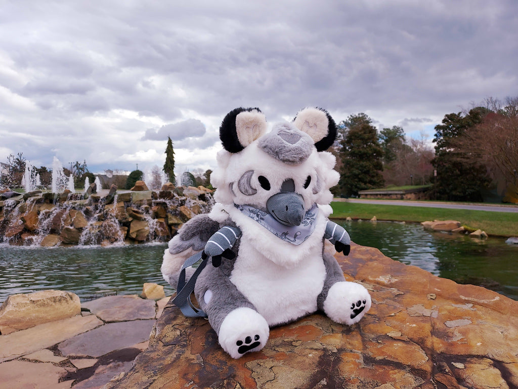 Plush backpack of Seasame the grey and white gryphon sitting on the rocks by a fountain