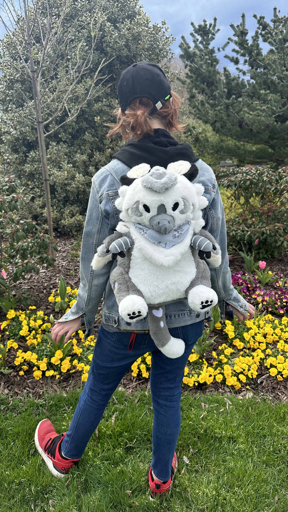 Person standing by some yellow flowers while wearing a plush backpack of Sesame the grey and white gryphon