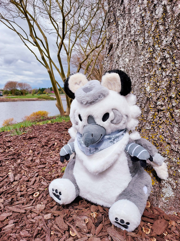 Plush backpack of Sesame the grey and white gryphon sitting on some mulch by a tree