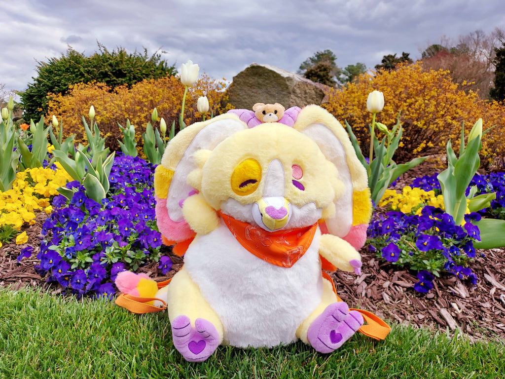 Plush backpack of Charmy the yellow dragon sitting amongst purple and white flowers