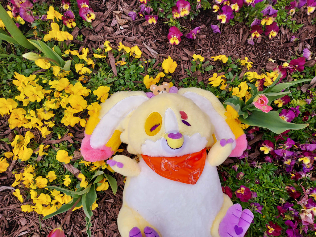 Plush backpack of Charmy the yellow dragon lying in a garden of yellow and purple flowers