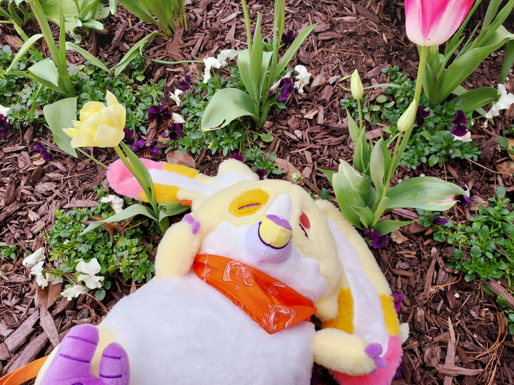 Plush backpack of Charmy the yellow dragon lying next to a pink tulip