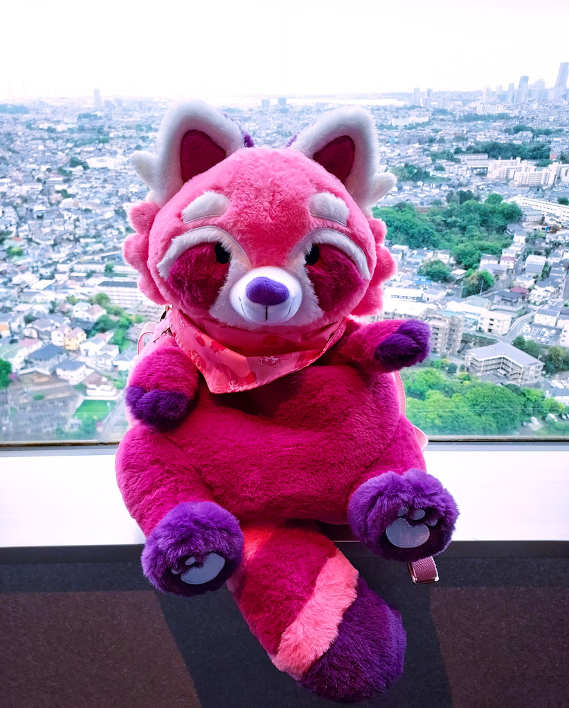 Plush backpack of Raspberry the red panda sitting in a window overlooking a city