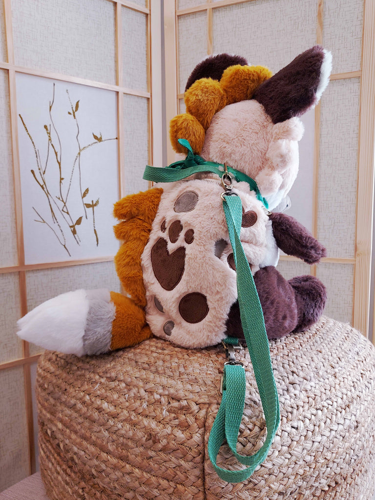 Back view of a plush backpack of Chocochip the hyena wearing a green bandana, with green backpack straps