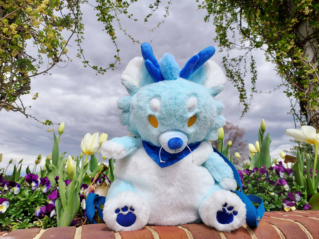 Plush backpack of Cloud the blue dragon sitting on a wall near white tulips