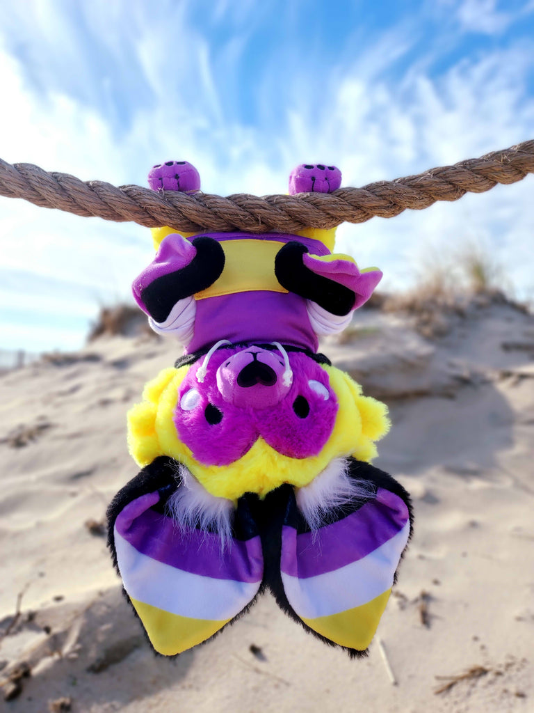 Plush toy of Butter the bat who is purple, yellow, white, and black, hanging upside down from a rope at the beach