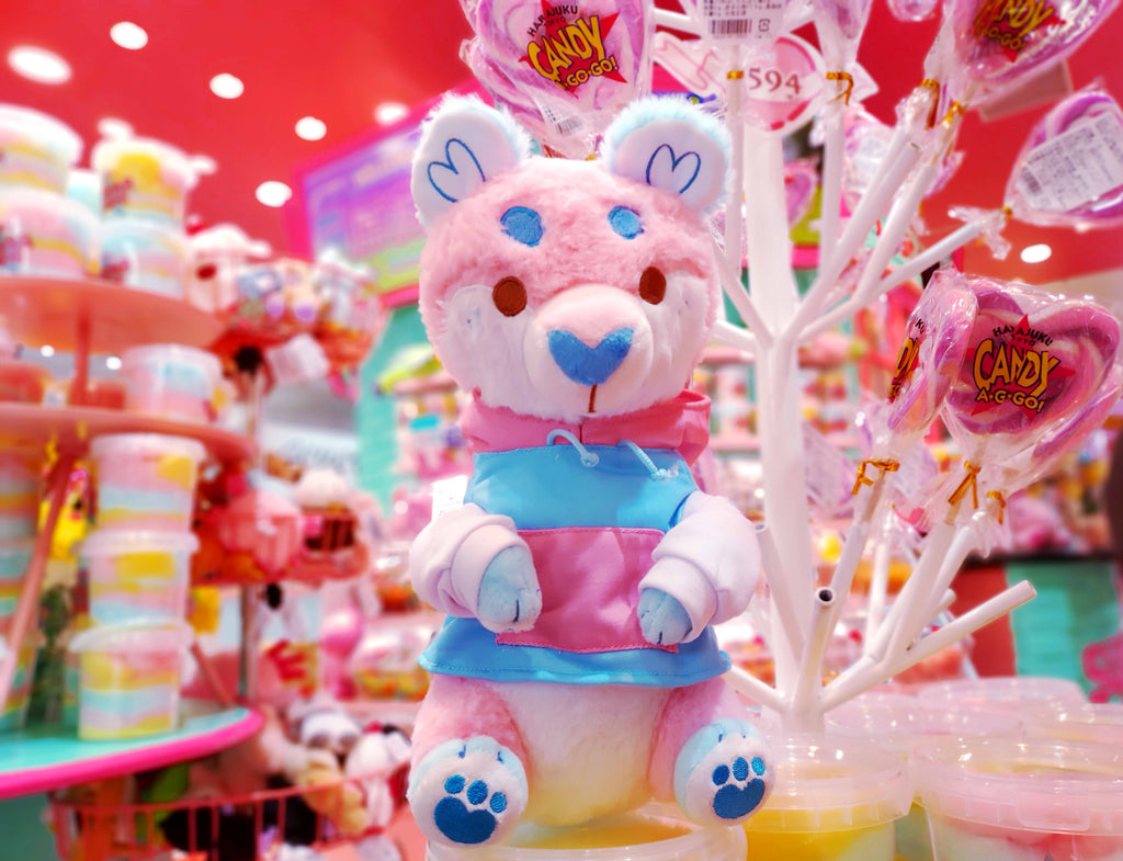 Plush toy of Cake Pop the otter who is pink, white, and blue and wearing a hoodie while at a pink candy shop's lollipop display