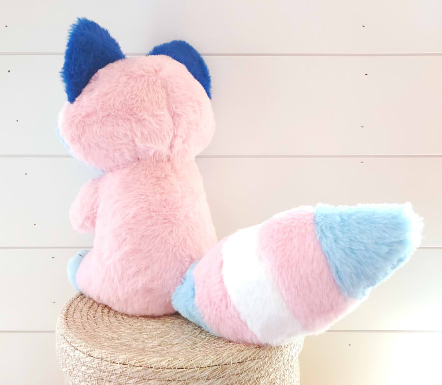 Back view of a plush toy of Egg the trans raccoon who is pink, blue, and white 