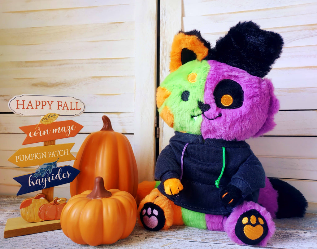 Snuggle Paws plush of THEM the monstrous abomination. They are half green and orange cat and half purple and black puppy. They are wearing a black hoodie and sitting next to pumpkins and a "Happy Fall" sign.