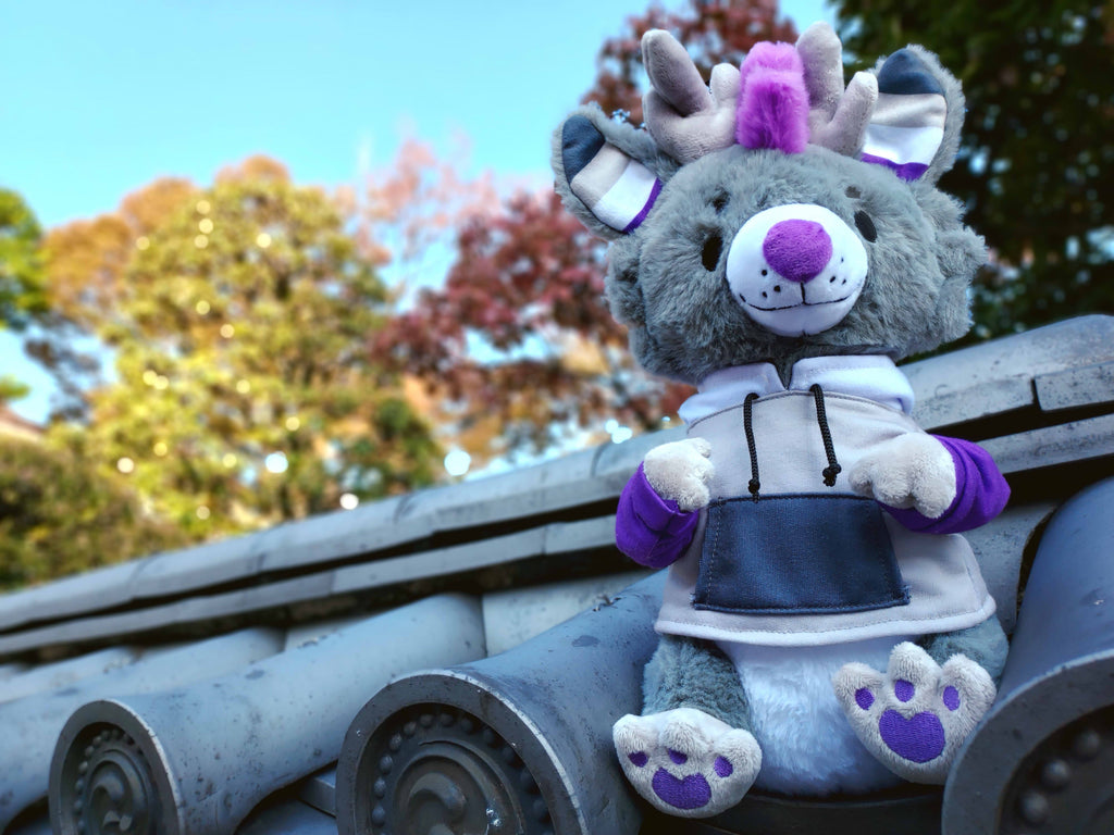 Plush toy of Marshmallow the dragon who is grey, white, black, and purple, sitting on the roof of a shrine in Asia