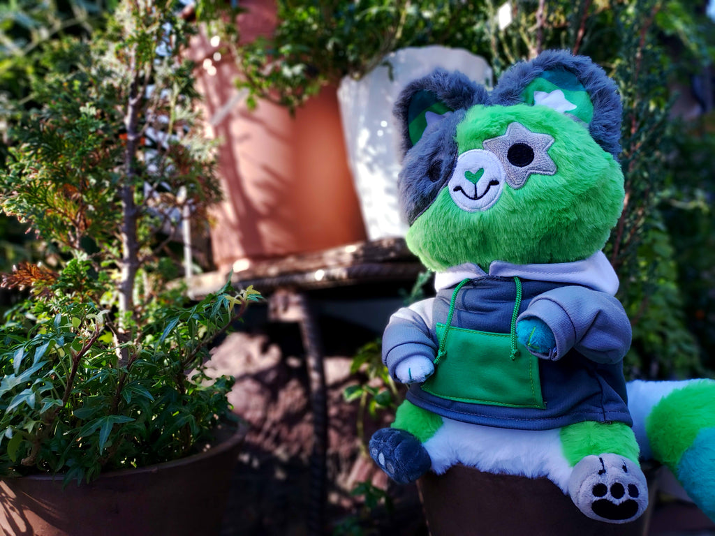 Plush toy of Matcha the cat who is green, black, white, and grey, wearing a hoodie and sitting amongst some plants