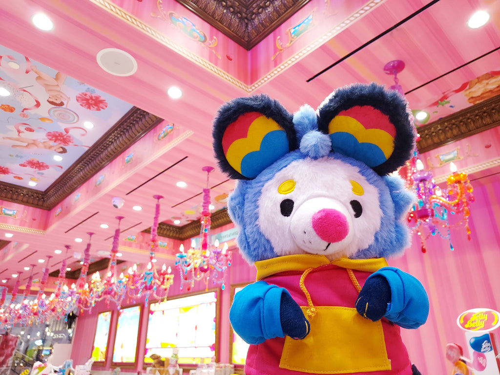 Plush toy of Pancake the possum who is blue, pink, and yellow, wearing a hoodie in a pink candy shop