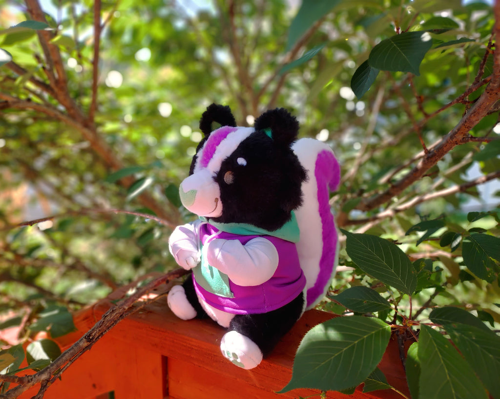 Plush toy of Pepper the green, purple, and white skunk, wearing a matching hoodie, sitting in a tree