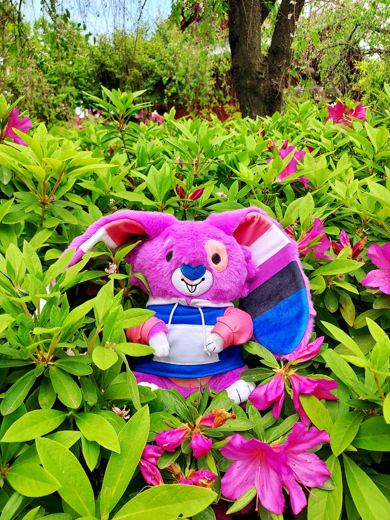 Plush toy of Bubblegum the purple, pink, black,white, and blue bunny wearing a colorful hoodie and relaxing in a garden with purple flowers