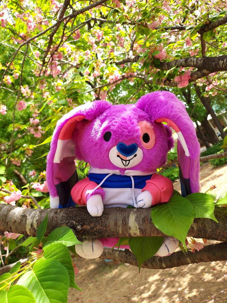 Plush toy of Bubblegum the purple, pink, black,white, and blue bunny wearing a colorful hoodie and relaxing in a tree