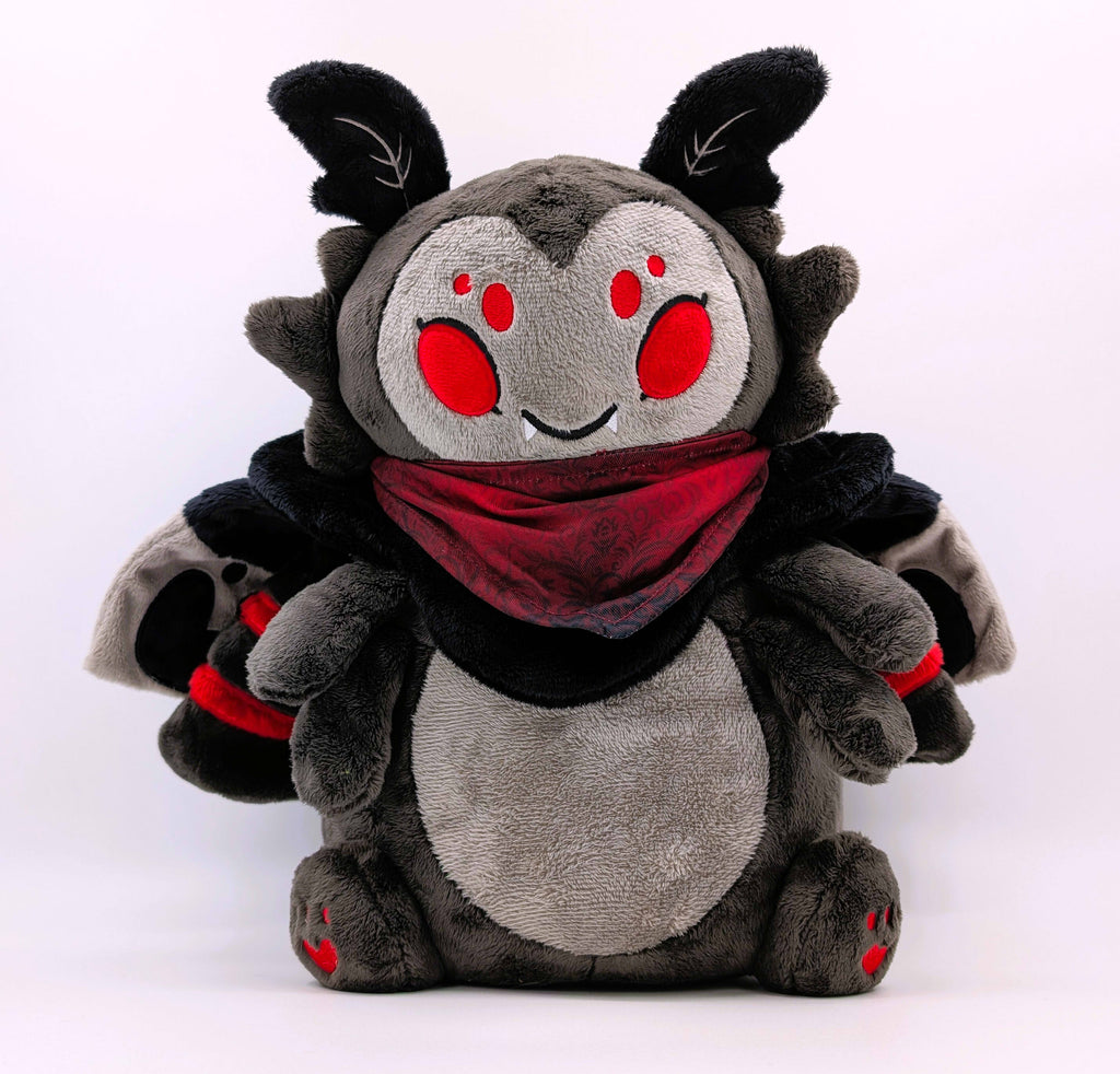 A plush toy of a black moth with red eyes wearing a red bandana. He is smiling and has fangs