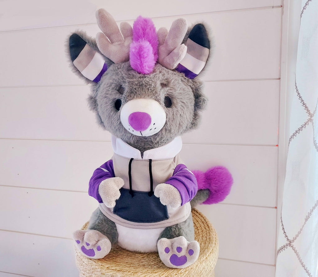 Plush of Marshmallow the dragon who is  black, purple, grey, and white, wearing a hoodie