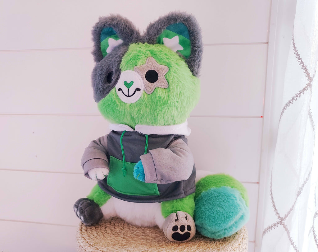 Plush toy of Matcha the cat who is green, black, white, and grey, wearing a hoodie