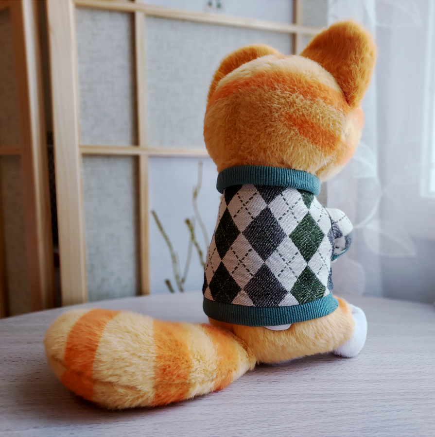 Back view of a plush toy of Cheese the orange cat wearing a green argyle sweater