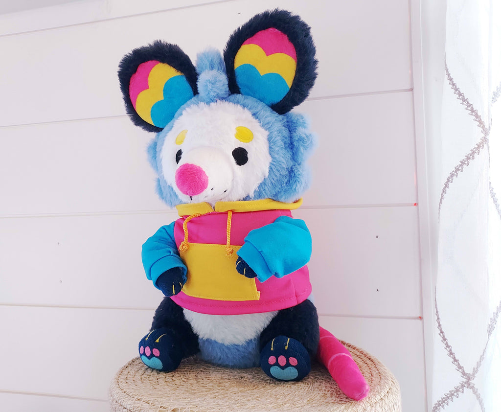 Plush toy of Pancake the possum who is blue, pink, and yellow, wearing a hoodie