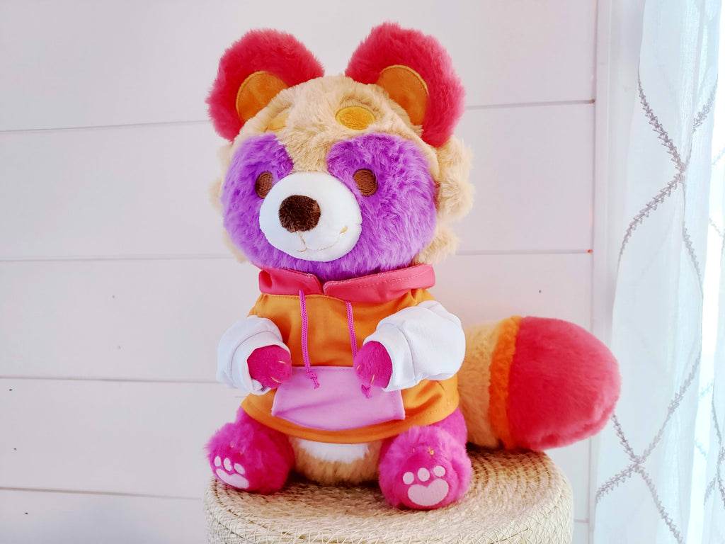 Plush toy of Sweet Potato the tanuki who is pink, purple, red, orange, and white wearing a hoodie 