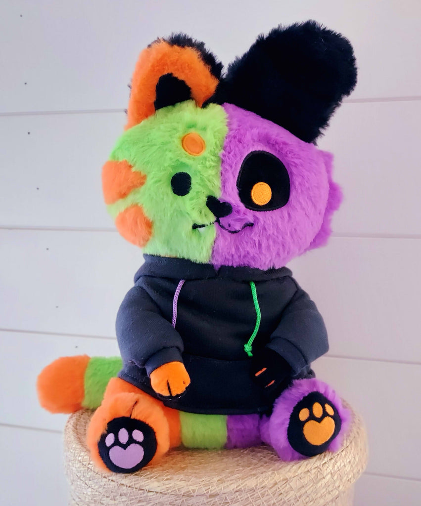 Snuggle Paws plush of THEM the monstrous abomination. They are half green and orange cat and half purple and black puppy. They are wearing a black hoodie.