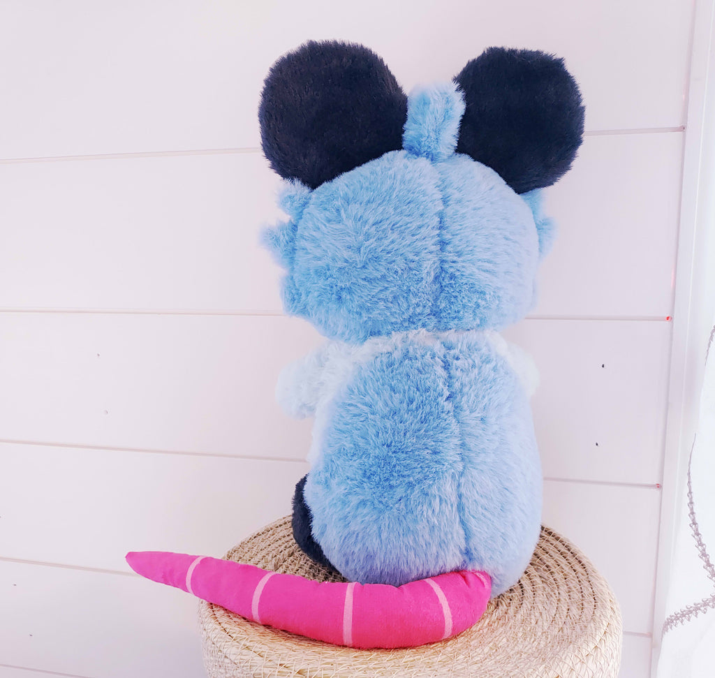 Back view of a plush toy of Pancake the possum who is blue, pink, and yellow