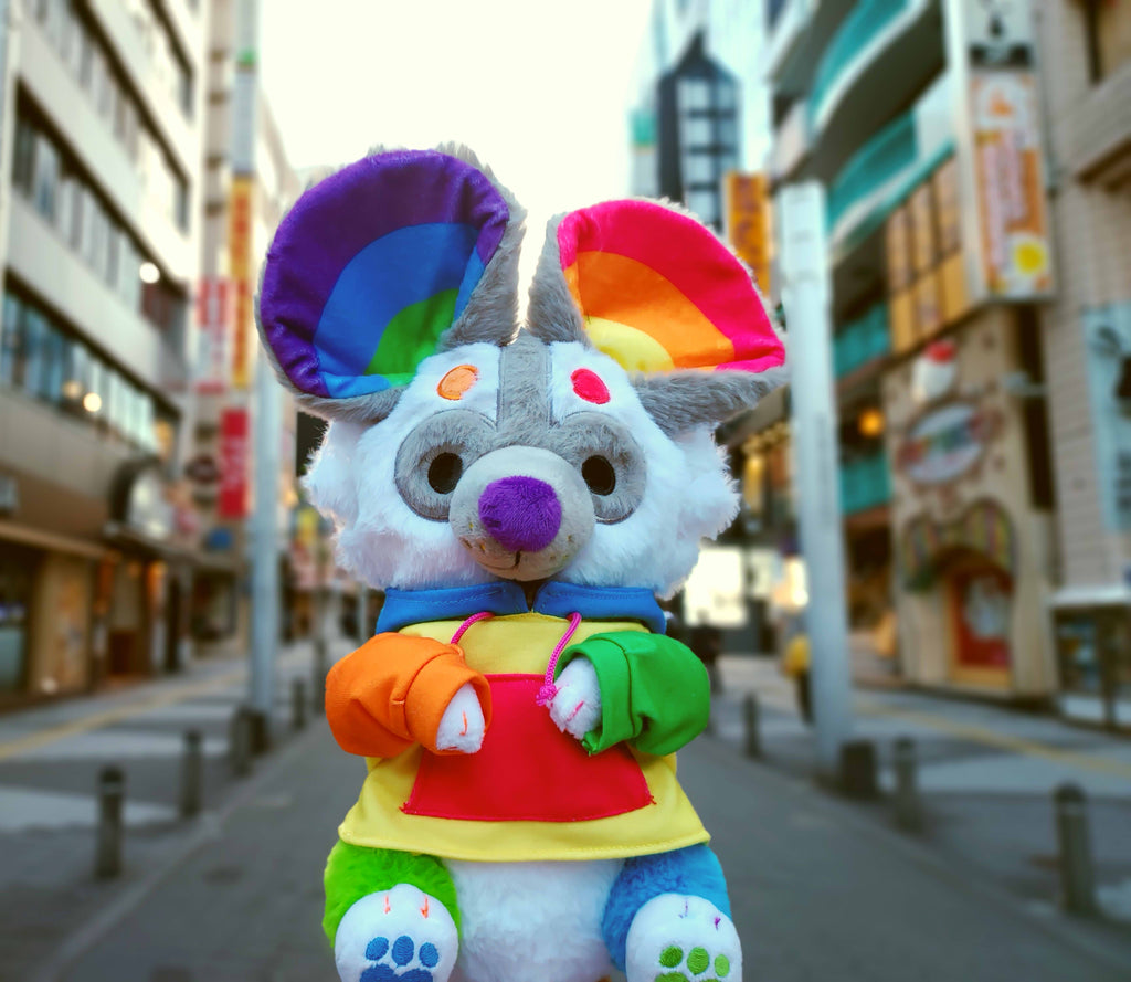 Plush toy of Sprinkles the rainbow painted dog wearing a rainbow hoodie on a city street