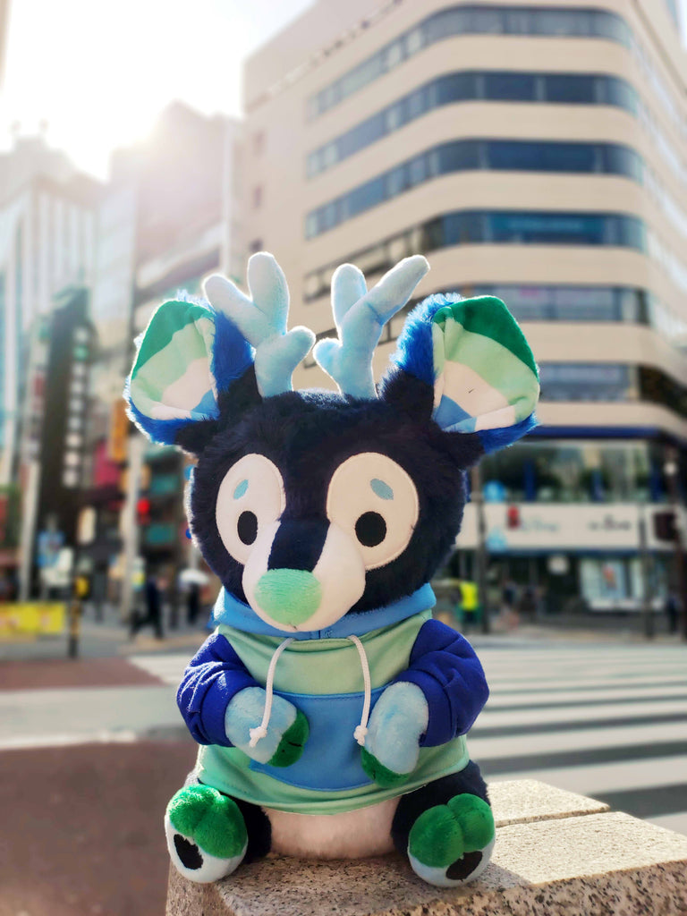 Plush toy of Smoothie the blue and green stag sitting on a city street