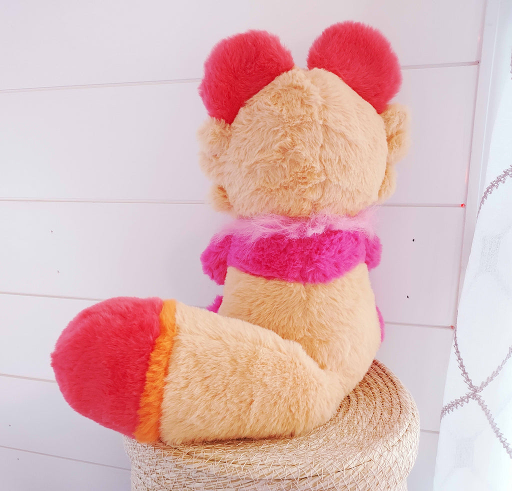 Back view of a plush toy of Sweet Potato the tanuki who is pink, purple, red, and orange